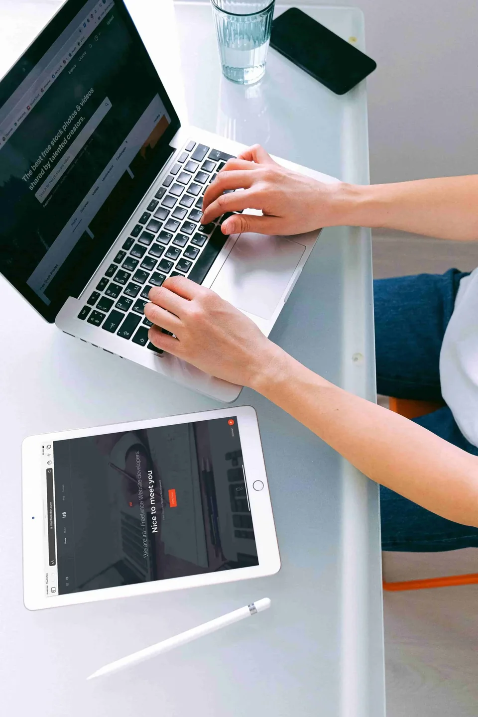 Hands typing on a laptop beside a tablet showing a website on a bright desk